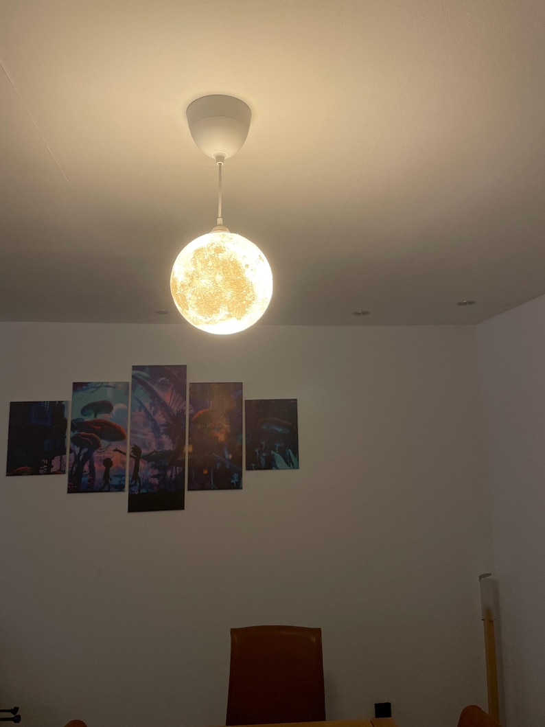 Eine kugelförmige Pendelleuchte mit einer detaillierten, strukturierten Oberfläche, die das Aussehen des Mondes nachahmt und ein warmes, leuchtendes Licht abgibt. Die Lampe hängt an einem weißen Kabel mit passendem weißen Deckenbaldachin.