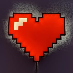May include: A pixelated red heart-shaped wall light with a black border. The heart has a white square in the upper left corner. The light is plugged into an electrical outlet.