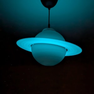 May include: A spherical pendant light fixture shaped like the planet Saturn, featuring a glowing cyan light emanating from within. The lamp includes a central globe and a surrounding ring structure, suspended from a dark ceiling mount by a thin cord.