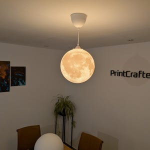 May include: A spherical pendant lamp featuring a detailed, textured surface that mimics the appearance of the moon. The lamp emits a warm, soft glow and is suspended from the ceiling by a white cord and a matching white circular ceiling mount.