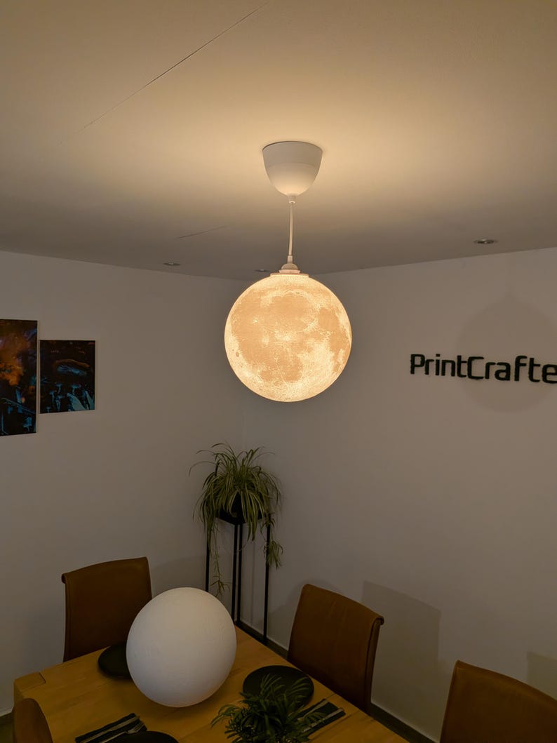 May include: A spherical pendant lamp featuring a detailed, textured surface that mimics the appearance of the moon. The lamp emits a warm, soft glow and is suspended from the ceiling by a white cord and a matching white circular ceiling mount.
