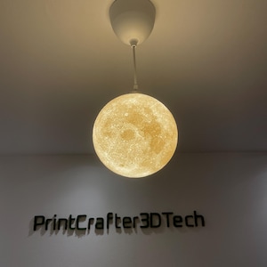Kugelförmige Mond-Lampe leuchtet warm-gelb und hängt an einem weißen Kabel von der Decke. Die 3D-gedruckte Oberfläche zeigt realistische Mondkrater. Darunter ein Wandlogo aus grünem Moos mit dem Schriftzug 'PrintCrafter3DTech