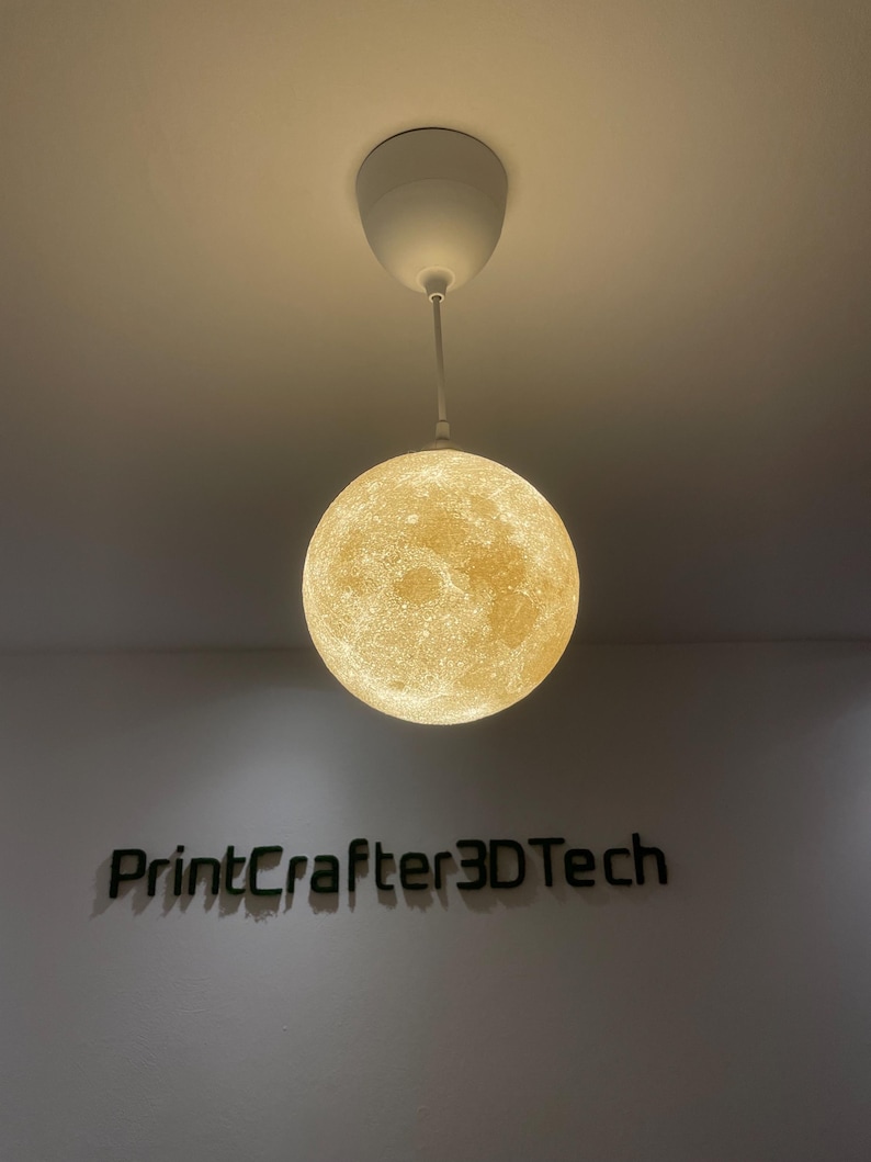 Kugelförmige Mond-Lampe leuchtet warm-gelb und hängt an einem weißen Kabel von der Decke. Die 3D-gedruckte Oberfläche zeigt realistische Mondkrater. Darunter ein Wandlogo aus grünem Moos mit dem Schriftzug 'PrintCrafter3DTech