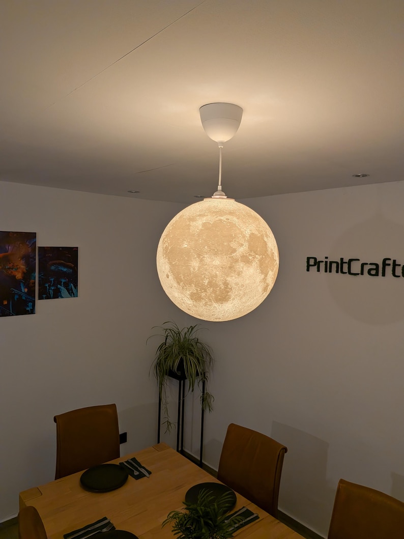 May include: A spherical pendant light fixture featuring a detailed, textured surface that mimics the appearance of the moon. The lamp emits a warm, soft glow and is suspended from the ceiling by a white cord and a matching white circular canopy.