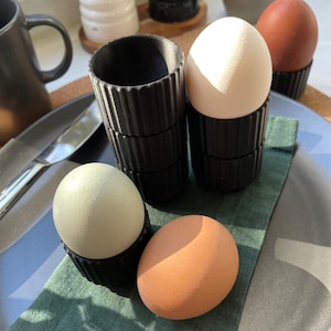 May include: Four eggs in black egg cups. Two eggs are white, one is light green, and one is brown. The egg cups are stacked on top of each other.