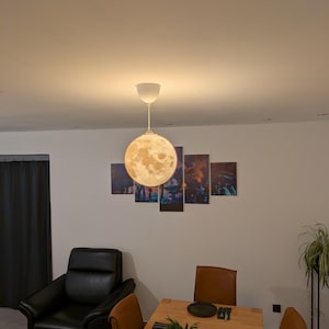 Könnte beinhalten: Eine kugelförmige Pendelleuchte mit einer detaillierten, strukturierten Oberfläche, die das Aussehen des Mondes nachahmt. Die Lampe strahlt ein warmes, sanftes Licht aus und ist mit einem dünnen weißen Kabel und einem weißen Baldachin befestigt.