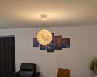 May include: A spherical pendant light fixture featuring a detailed, textured surface that mimics the appearance of the moon. The lamp emits a warm, soft glow and is suspended from the ceiling by a thin white cord and a matching white canopy.