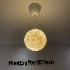 May include: A spherical pendant light fixture featuring a textured, cratered surface resembling the moon, suspended from a white ceiling canopy by a thin white cord. Below the light, the text PrintCrafter3DTech is mounted on the wall.