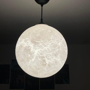 Puede incluir: Una lámpara colgante esférica que se asemeja a la luna. La luz tiene una superficie texturizada con un diseño similar a un cráter y cuelga de un cable negro y un soporte de techo. Emite un brillo suave y ambiental.