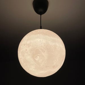 Puede incluir: Una lámpara colgante esférica de color blanquecino que se asemeja a la luna. La luz tiene una superficie texturizada con cráteres y un cable y soporte de techo negros. La luz está suspendida sobre un fondo oscuro.