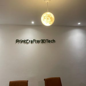 Eine kugelförmige, mondartige Pendelleuchte mit strukturierter Oberfläche hängt von der Decke. Darunter befindet sich der Schriftzug "PrintCrafter3DTech" an einer schlichten weißen Wand, platziert über zwei braunen Stühlen und einem Holztisch.