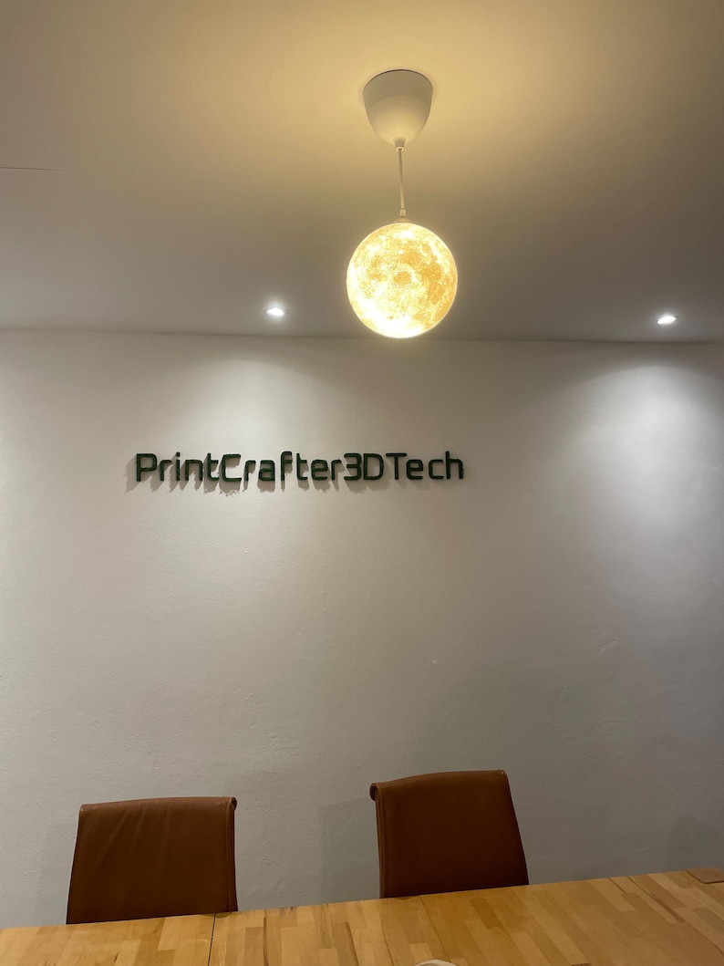Eine kugelförmige, mondartige Pendelleuchte mit strukturierter Oberfläche hängt von der Decke. Darunter befindet sich der Schriftzug "PrintCrafter3DTech" an einer schlichten weißen Wand, platziert über zwei braunen Stühlen und einem Holztisch.