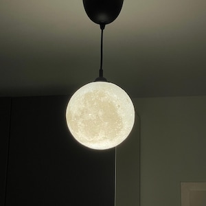 Eine kugelförmige Pendelleuchte mit einer detaillierten, strukturierten Oberfläche, die das Aussehen des Mondes nachahmt. Die Kugel wird von innen beleuchtet, strahlt ein sanftes Licht aus und hängt an einem schwarzen Kabel an einem schwarzen Deckenbaldachin.