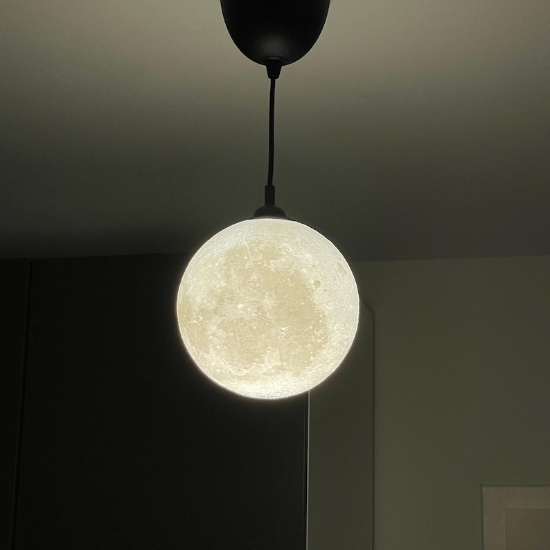 Eine kugelförmige Pendelleuchte mit einer detaillierten, strukturierten Oberfläche, die das Aussehen des Mondes nachahmt. Die Kugel wird von innen beleuchtet, strahlt ein sanftes Licht aus und hängt an einem schwarzen Kabel an einem schwarzen Deckenbaldachin.