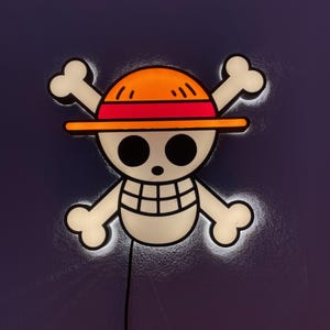 Könnte beinhalten: Leuchtende Wanddekoration mit dem Jolly Roger-Symbol aus der Anime-Serie One Piece. Das Design zeigt einen weißen Totenkopf mit gekreuzten Knochen, einen Strohhut mit orangefarbenen und roten Akzenten und ein schwarzes Netzkabel.
