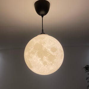 May include: A spherical pendant light fixture featuring a detailed, textured surface that mimics the appearance of the moon. The globe is illuminated from within, displaying craters and lunar topography in shades of white and gray, suspended by a black cord.