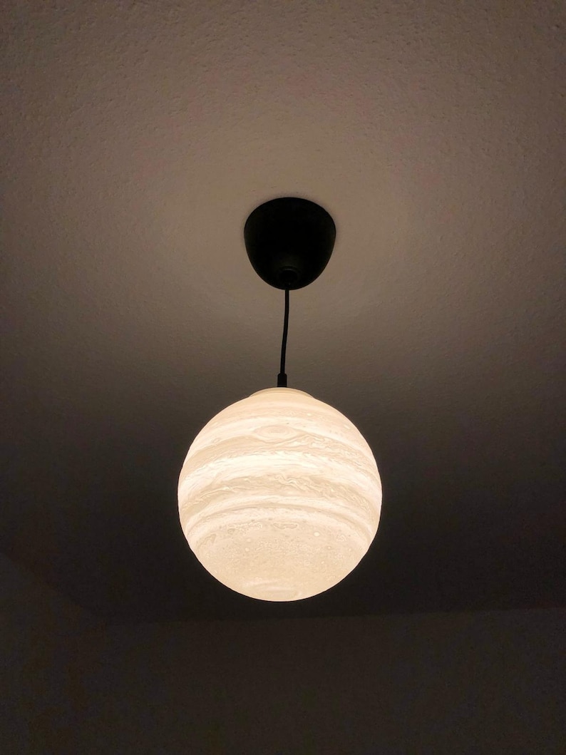 Eine kugelförmige Pendelleuchte mit einer strukturierten, cremeweißen Oberfläche und horizontalen Bändern Sie stellt den Jupiter dar, die an das Aussehen eines Planeten erinnern. Die Kugel hängt an einem dünnen schwarzen Kabel an einem schwarzen, runden Deckenbaldachin.