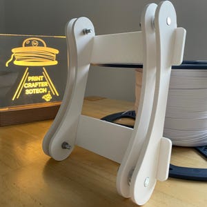 Puede incluir: Un soporte blanco para bobinas de filamento de impresora 3D. El soporte está hecho de madera blanca y tiene un marco rectangular. El logotipo "PRINT CRAFTER 3DTECH" es visible en el fondo. El soporte está diseñado para sujetar una bobina de filamento.