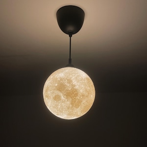 Mond-Lampe mit warmem, goldenem Licht hängt an einem schwarzen Kabel und schwarzer Deckenhalterung. Die Oberfläche zeigt detaillierte, kraterartige Strukturen, die im Dunkeln besonders leuchten.