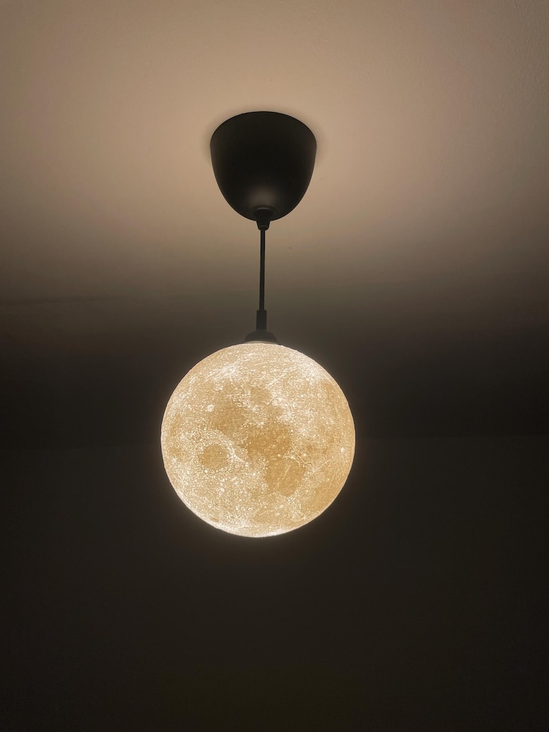 Mond-Lampe mit warmem, goldenem Licht hängt an einem schwarzen Kabel und schwarzer Deckenhalterung. Die Oberfläche zeigt detaillierte, kraterartige Strukturen, die im Dunkeln besonders leuchten.