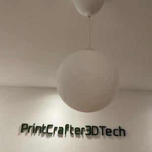 Runde Mond-Lampe hängt an einem weißen Kabel von der Decke, ausgeschaltet. Die Oberfläche zeigt eine strukturierte, kraterartige Textur in Weiß. Darunter der Schriftzug 'PrintCrafter3DTech' aus grünem Moos an der Wand.