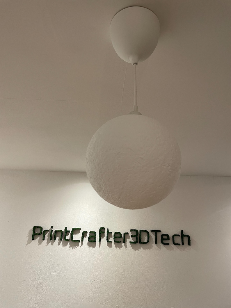 Runde Mond-Lampe hängt an einem weißen Kabel von der Decke, ausgeschaltet. Die Oberfläche zeigt eine strukturierte, kraterartige Textur in Weiß. Darunter der Schriftzug 'PrintCrafter3DTech' aus grünem Moos an der Wand.