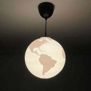 Może przedstawiać: Lampa wisząca w kształcie kuli ziemskiej. Biała kula z mapą świata w jasnoróżowym kolorze. Lampa zawieszona jest na czarnym uchwycie sufitowym za pomocą czarnego kabla.