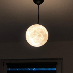 Eine kugelförmige Pendelleuchte mit einer detaillierten, strukturierten Oberfläche, die das Aussehen des Mondes nachahmt. Die Lampe ist an einem schwarzen Kabel an einem passenden schwarzen, runden Deckenbaldachin befestigt.