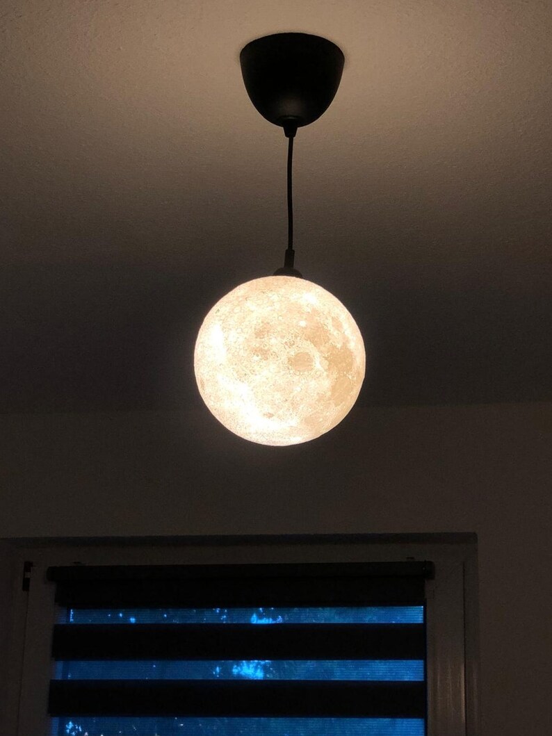 Eine kugelförmige Pendelleuchte mit einer detaillierten, strukturierten Oberfläche, die das Aussehen des Mondes nachahmt. Die Lampe ist an einem schwarzen Kabel an einem passenden schwarzen, runden Deckenbaldachin befestigt.