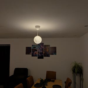 May include: A spherical moon-shaped pendant light fixture hangs from the ceiling above a wooden dining table. The table is set with black plates, a small potted plant, and a candle. A five-panel wall art piece is mounted on the wall behind the table.