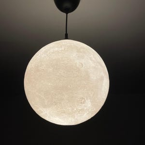 Może przedstawiać: Sferyczna, biała lampa wisząca przypominająca księżyc. Światło ma teksturowaną powierzchnię z kraterami oraz czarny przewód i mocowanie sufitowe. Światło jest zawieszone na ciemnym tle.