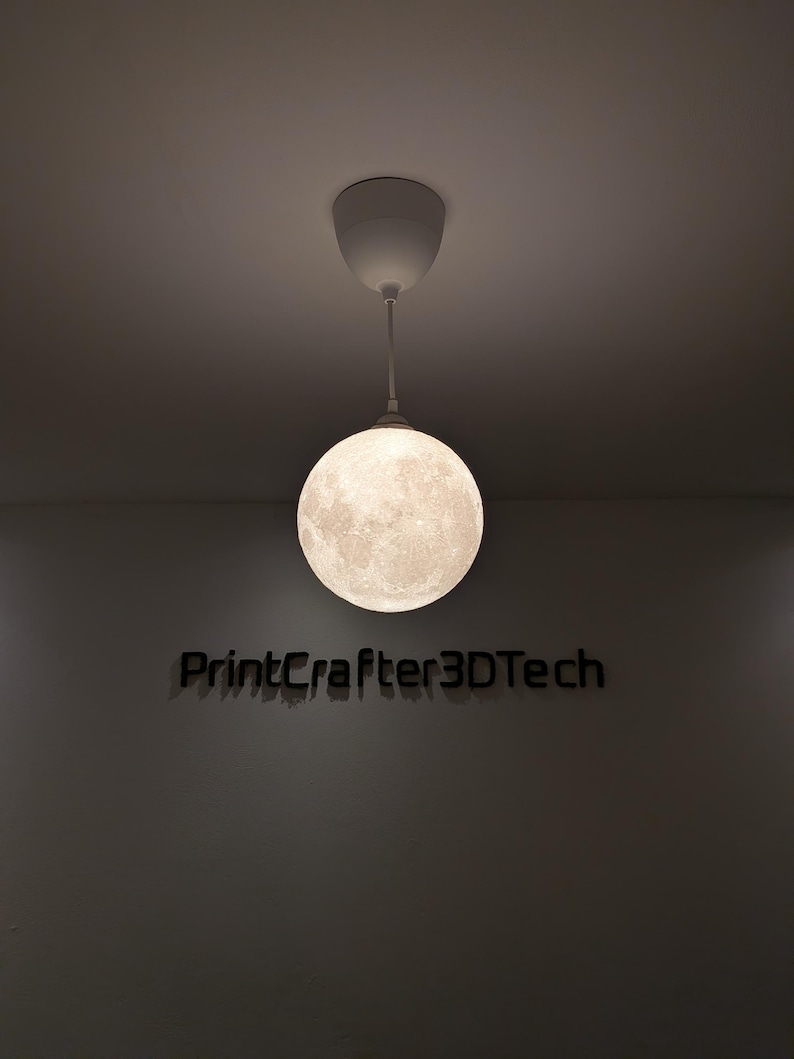 Eine kugelförmige Pendelleuchte mit einer detaillierten, strukturierten Oberfläche, die dem Mond ähnelt und an einer weißen Deckenhalterung hängt. Unter der beleuchteten Mondlampe ist der Text PrintCrafter3DTech in dunkler, fetter Schrift zu sehen.