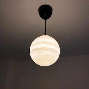 May include: A spherical pendant light fixture featuring a white, textured, marbled surface that glows when illuminated. The globe is suspended from a thin black cord attached to a black circular ceiling canopy mounted on a plain ceiling.
