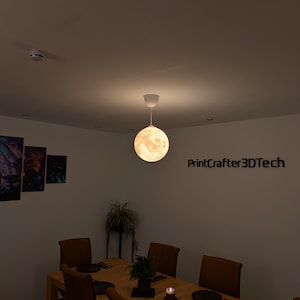 Eine kugelförmige, mondförmige Pendelleuchte hängt von der Decke und strahlt ein warmes Licht aus. Die Oberfläche der Leuchte zeigt detaillierte Mondkrater und Texturen. Der Text PrintCrafter3DTech ist an der Wand neben der Leuchte sichtbar.