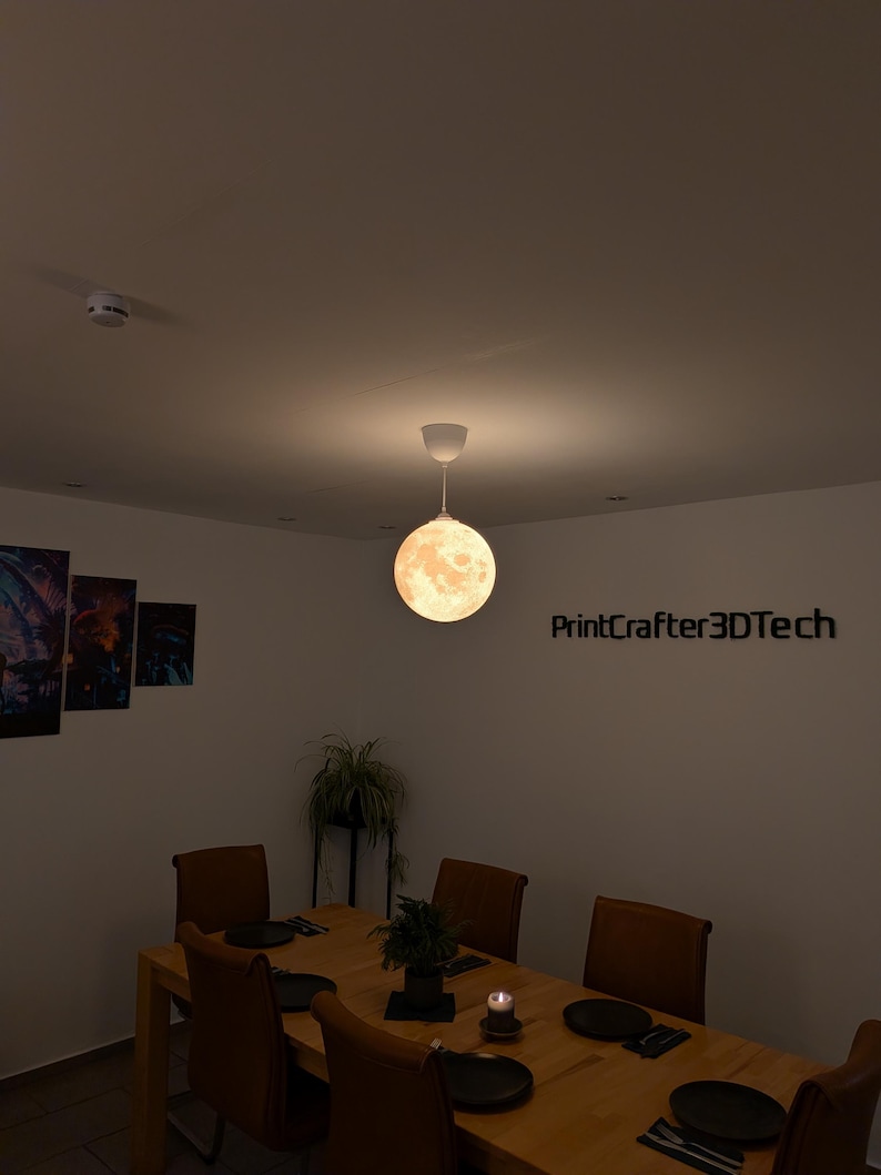 Eine kugelförmige, mondförmige Pendelleuchte hängt von der Decke und strahlt ein warmes Licht aus. Die Oberfläche der Leuchte zeigt detaillierte Mondkrater und Texturen. Der Text PrintCrafter3DTech ist an der Wand neben der Leuchte sichtbar.