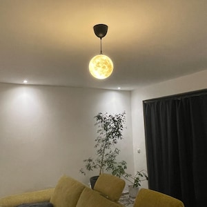 Kleine, leuchtende Mond-Lampe hängt an schwarzem Kabel von einer Zimmerdecke über einem gelben Sofa. Im Hintergrund eine Zimmerpflanze und dunkle Vorhänge, die eine gemütliche Wohnzimmeratmosphäre zeigen.