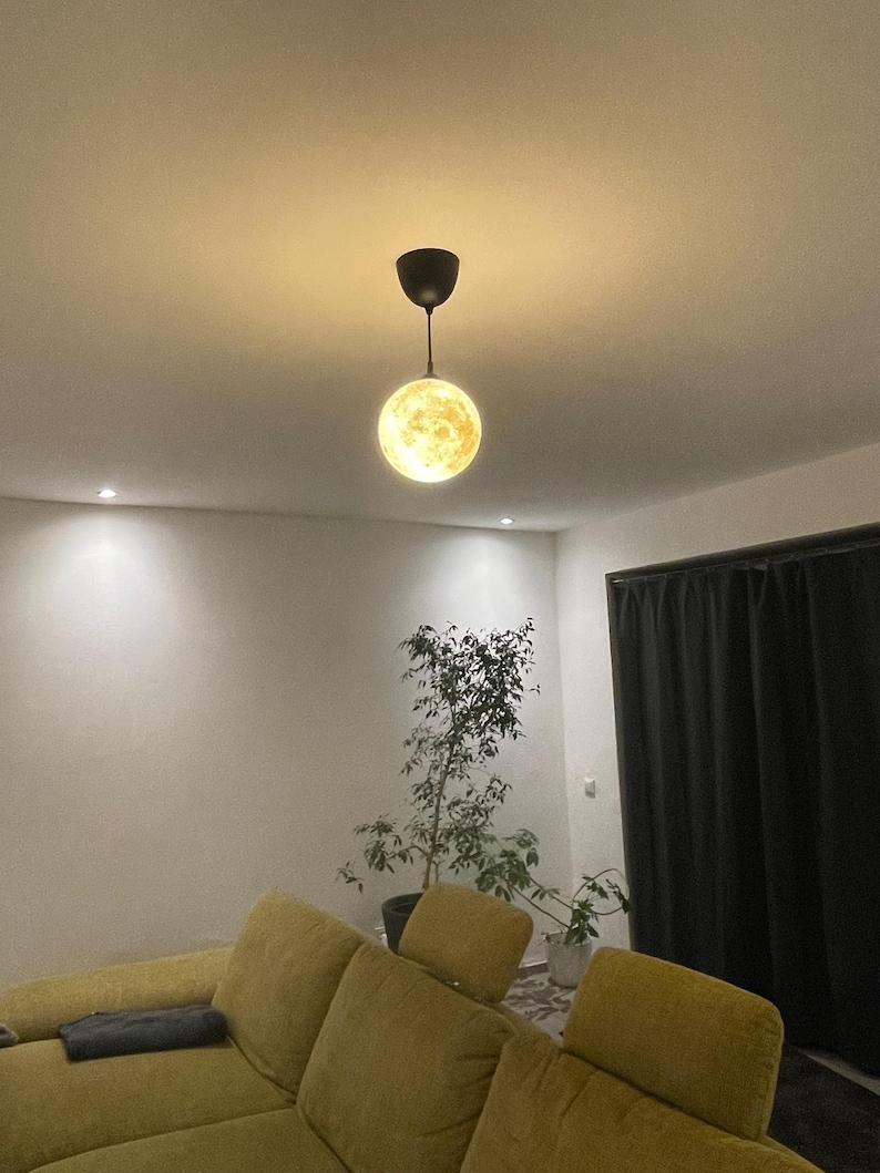 Kleine, leuchtende Mond-Lampe hängt an schwarzem Kabel von einer Zimmerdecke über einem gelben Sofa. Im Hintergrund eine Zimmerpflanze und dunkle Vorhänge, die eine gemütliche Wohnzimmeratmosphäre zeigen.