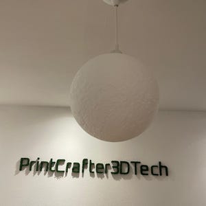 May include: A white spherical pendant light with a textured surface hangs from a white cord. Below the light, the text PrintCrafter3DTech is displayed in dark green, three-dimensional lettering mounted directly onto the wall surface.