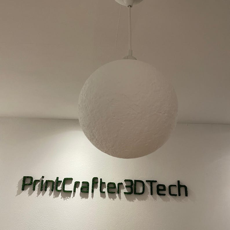 May include: A white spherical pendant light with a textured surface hangs from a white cord. Below the light, the text PrintCrafter3DTech is displayed in dark green, three-dimensional lettering mounted directly onto the wall surface.