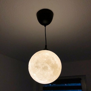 Eine kugelförmige Pendelleuchte, die mit einem schwarzen Kabel und Baldachin an der Decke befestigt ist. Der Globus hat eine strukturierte, cremeweiße Oberfläche, die das kraterartige Aussehen des Mondes nachahmt und leuchtet.