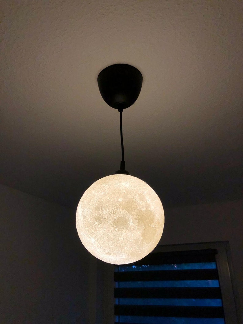 Eine kugelförmige Pendelleuchte, die mit einem schwarzen Kabel und Baldachin an der Decke befestigt ist. Der Globus hat eine strukturierte, cremeweiße Oberfläche, die das kraterartige Aussehen des Mondes nachahmt und leuchtet.