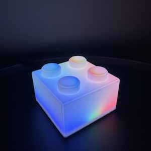 Könnte beinhalten: Eine quadratische, halbtransparente weiße Lampe in Form eines Bausteins mit vier runden Noppen auf der Oberseite. Die Lampe strahlt ein sanftes, mehrfarbiges Licht aus, mit blauem Licht auf der linken Seite und einem Farbverlauf in Rot und Grün rechts.