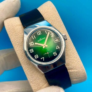 May include: A vintage Morex wristwatch featuring a round dial with a green gradient finish, white Arabic numerals, and a date window at the three o'clock position. The watch has a silver-tone metal case and a smooth black leather strap.
