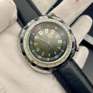 Puede incluir: Reloj de pulsera vintage con una correa de cuero negro. La esfera del reloj es de color verde oscuro con detalles en blanco y dorado, incluyendo una escala taquimétrica e indicadores de ciudades del mundo. El reloj tiene una corona dorada y un bisel negro.