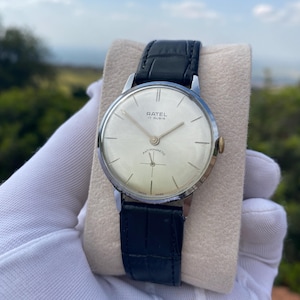 Op de afbeelding: Een vintage zilverkleurig RATEL horloge met een crèmekleurige wijzerplaat en een zwarte leren band. De wijzerplaat toont de tekst "RATEL 17 RUBIS ANTIMAGNETIC SWISS MADE". Het horloge wordt op een beige oppervlak gehouden.