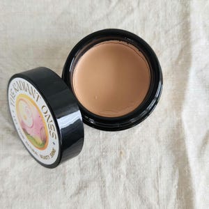 May include: A round black cosmetic jar containing beige cream, with a detached black lid featuring a circular label that reads THE RADIANT ONES + LET YOUR NATURAL BEAUTY SHINE and displays a stylized illustration of a face in pink and yellow.