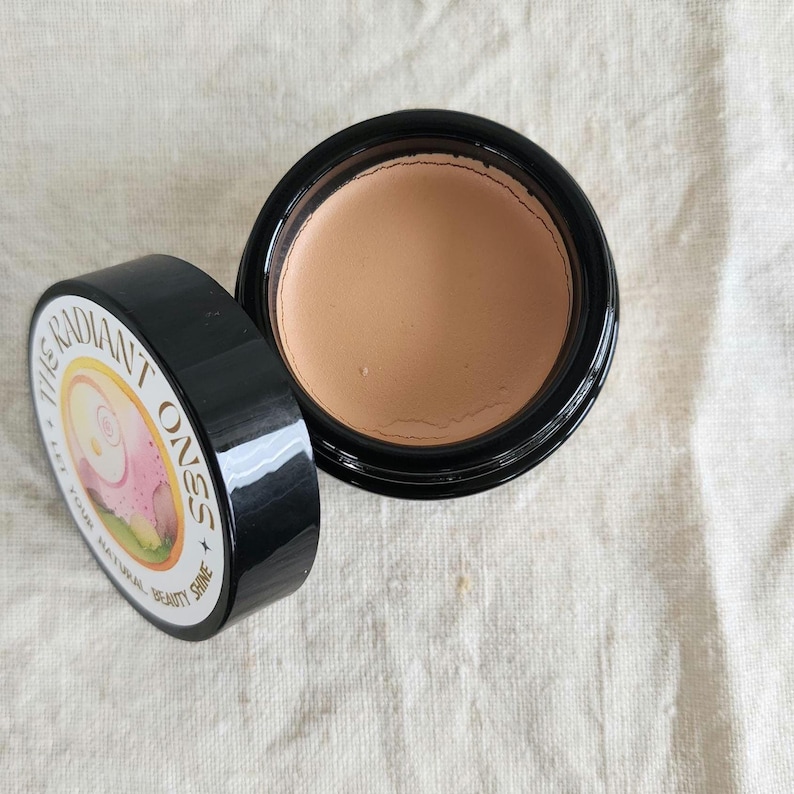 May include: A round black cosmetic jar containing beige cream, with a detached black lid featuring a circular label that reads THE RADIANT ONES + LET YOUR NATURAL BEAUTY SHINE and displays a stylized illustration of a face in pink and yellow.