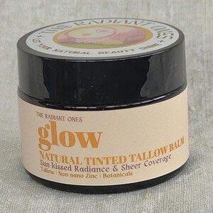 May include: A round black jar of The Radiant Ones glow natural tinted tallow balm. The beige label features the text: The Radiant Ones, glow, Natural Tinted Tallow Balm, Sun-kissed Radiance & Sheer Coverage, Tallow, Non-nano Zinc, Botanicals.