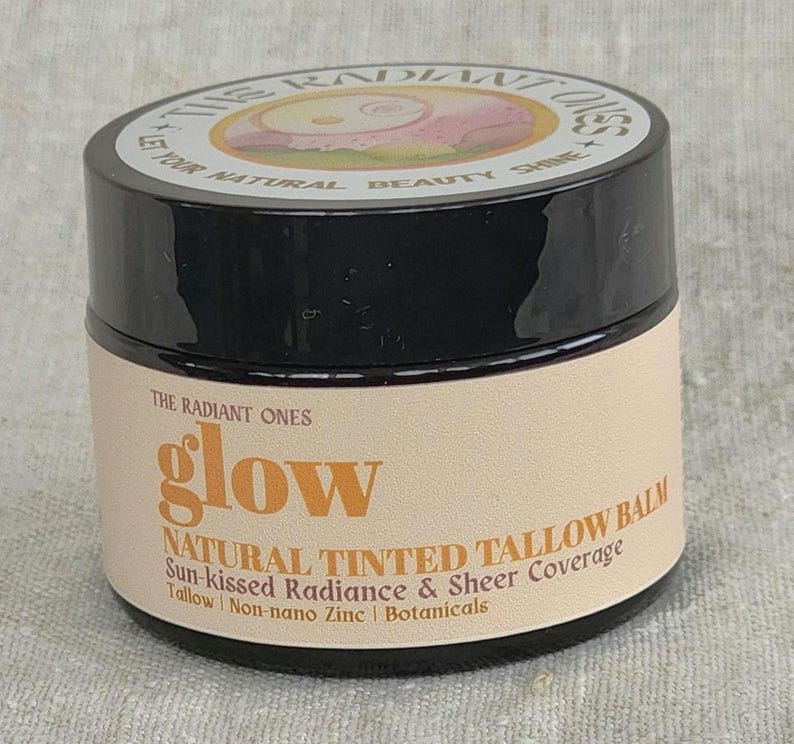 May include: A round black jar of The Radiant Ones glow natural tinted tallow balm. The beige label features the text: The Radiant Ones, glow, Natural Tinted Tallow Balm, Sun-kissed Radiance & Sheer Coverage, Tallow, Non-nano Zinc, Botanicals.