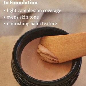 May include: A black jar containing a beige-toned cosmetic balm with a wooden spatula scooping a portion. Text reads: A Skin-Loving Alternative to Foundation, light complexion coverage, evens skin tone, nourishing balm texture.